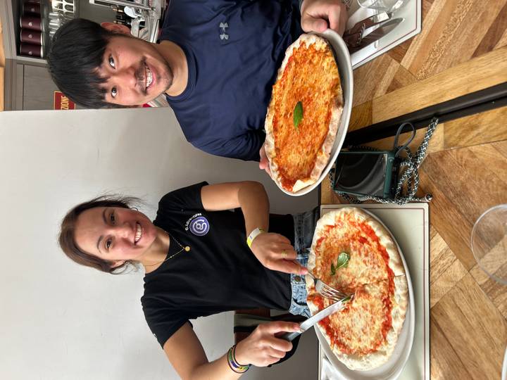 Two diners sit with whole Margherita pizzas on a wooden table, smiling for the camera.