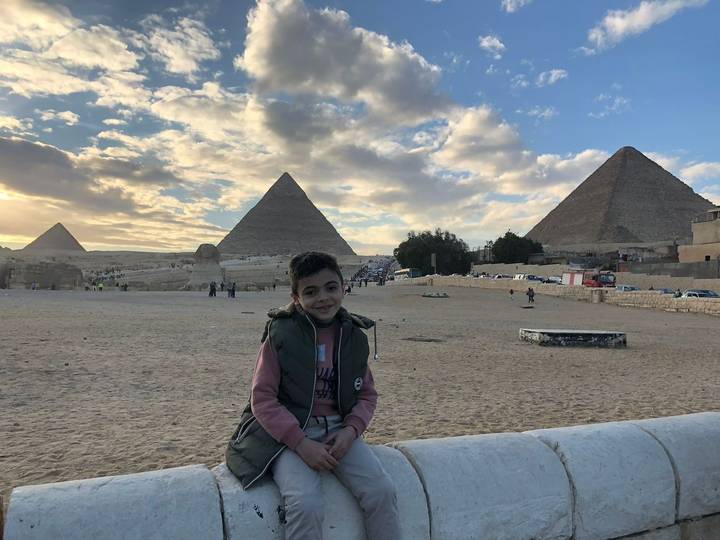 Smiling boy sits on a low wall with three pyramids silhouetted against a dramatic evening sky.