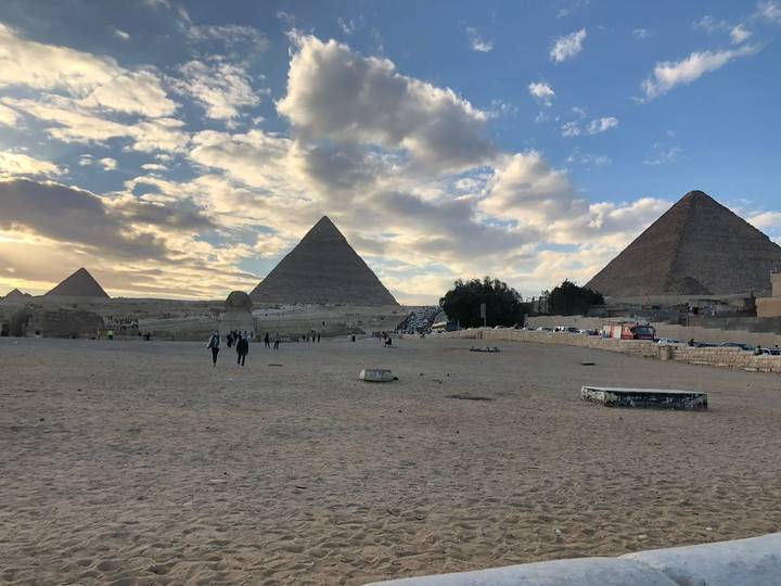 Wide desert scene with the three Pyramids of Giza rising behind a few visitors under a dramatic partly-cloudy sky.