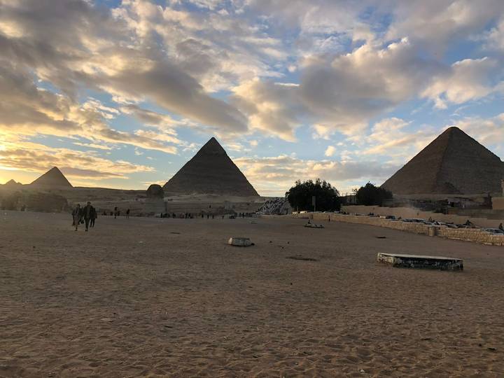 Sunny late-day view of the Giza pyramids with a few people silhouetted on the sandy plateau and glowing clouds overhead.