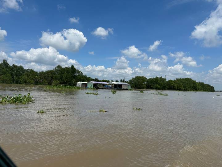 Wide muddy river scene with floating vegetation and a few stilted houses under a bright blue sky.