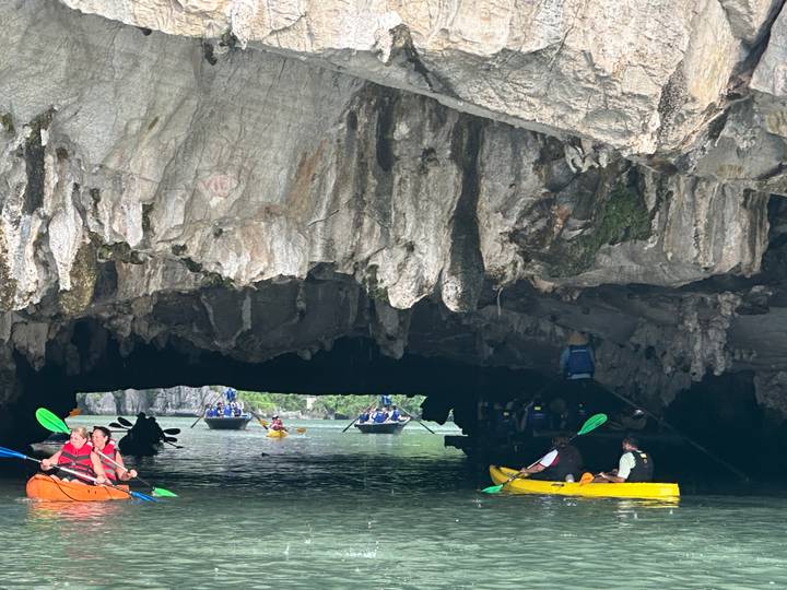 Kayakers paddle through emerald water toward a dramatic limestone cave entrance with stalactites.