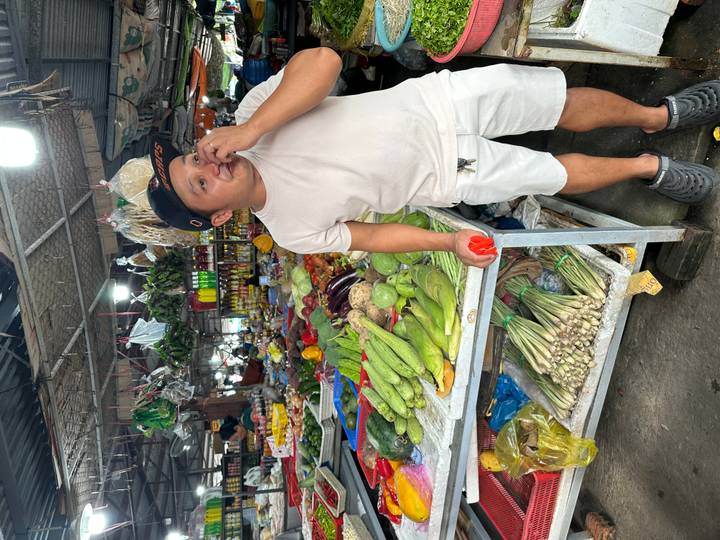 Market vendor stands beside a colourful stall piled high with fresh vegetables and herbs.