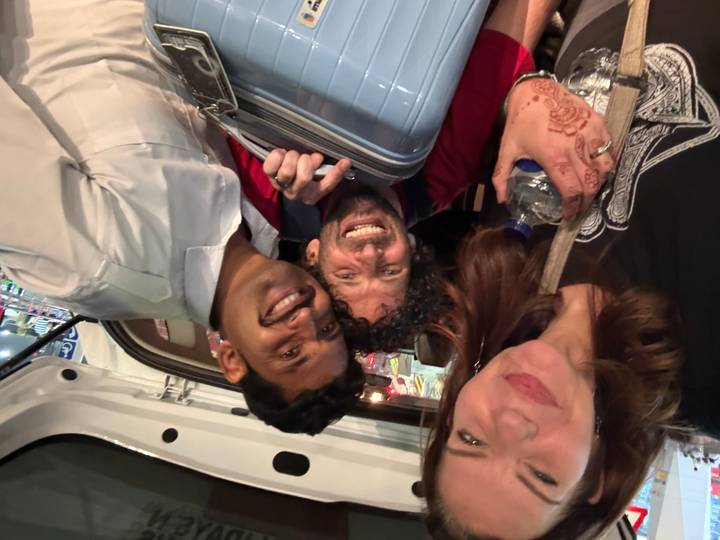 Night-time selfie of three travellers inside a vehicle, smiling while holding luggage.