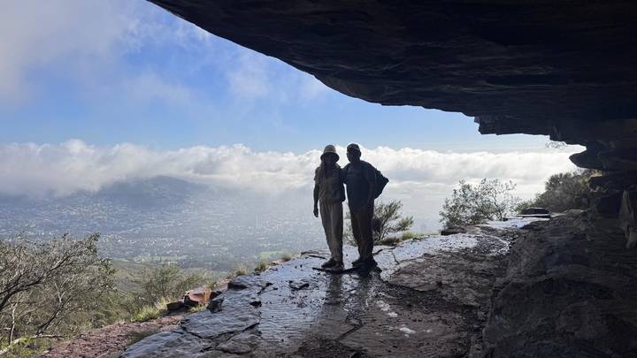 Silhouetted pair standing at the mouth of a rocky overhang, overlooking misty hills and a cloud-filled valley.