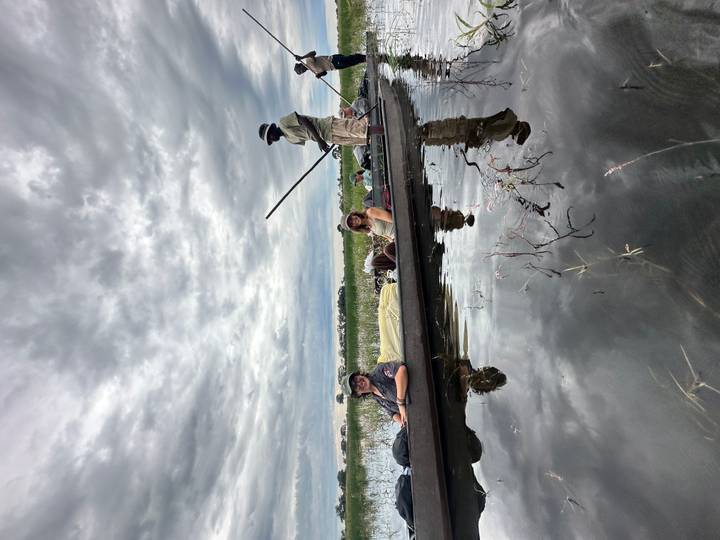 Tourists reclining in a dug-out canoe while a guide poles along a reflective wetland under dramatic cloudy skies.