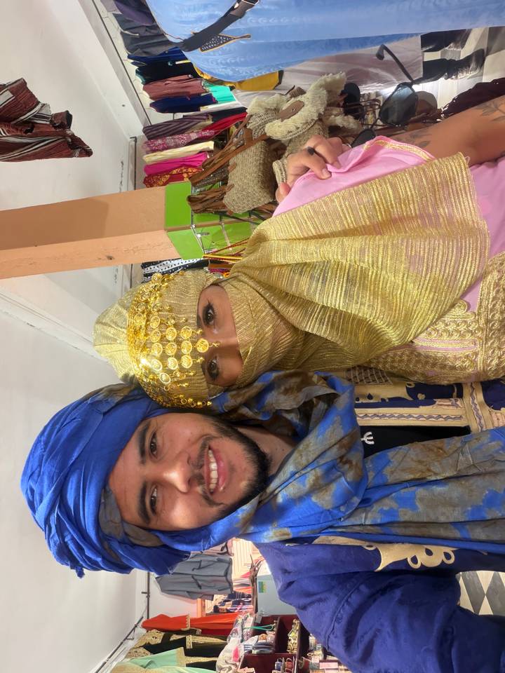 Close-up half-frame selfie of couple wearing golden and blue traditional headgear indoors.
