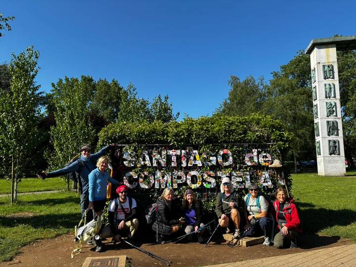 Pilgrim group poses by leafy Santiago de Compostela sign in sunny park.