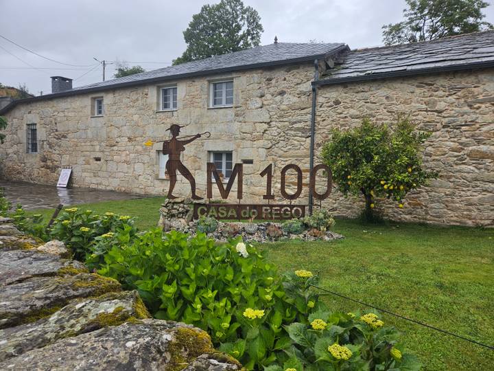 Sign for KM100 Casa do Rego on the Camino set in lush garden beside stone wall.