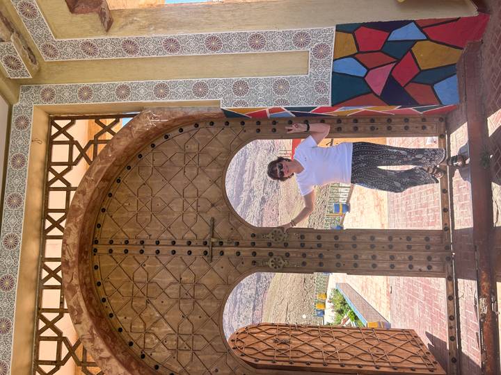 Traveller flashes peace sign while standing in decorative Moorish doorway with desert hills behind.