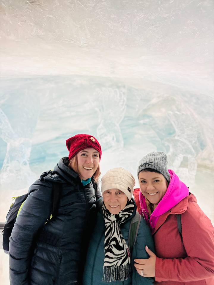 Three women in winter gear smiling inside an icy cave with carved walls