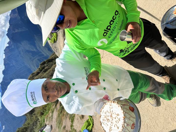 Trail chef in white uniform presents a cake while guide in bright green shirt smiles against Andean peaks