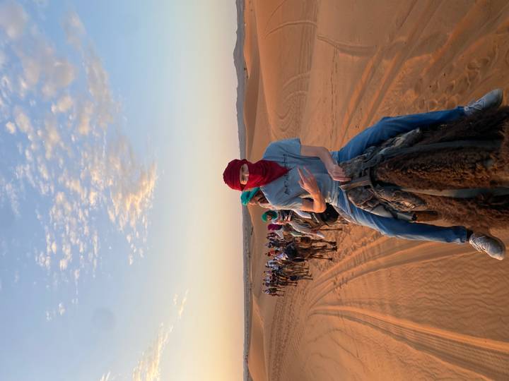 A long caravan of camel riders snakes across glowing orange dunes at sunrise beneath a pastel sky.