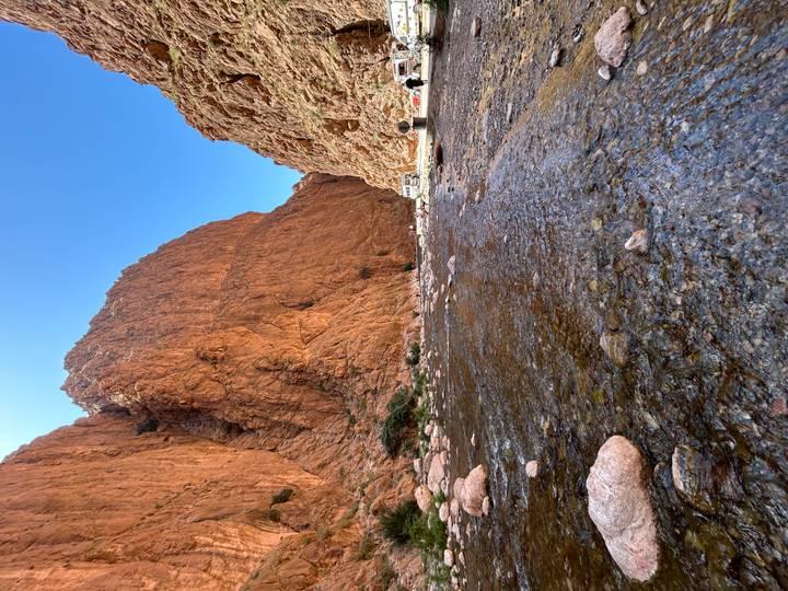 Clear stream flows over pebbles through a narrow red-rock canyon under a bright blue sky.