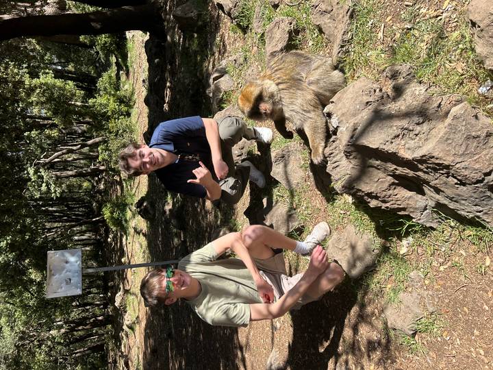 Two travellers crouch beside a relaxed Barbary macaque in a dappled forest clearing.