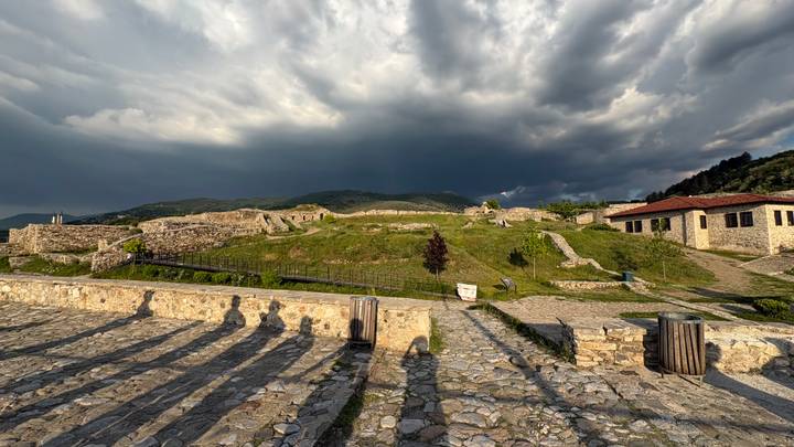Stone fortress ruins under dramatic storm clouds with long human shadows cast on cobbled ground.