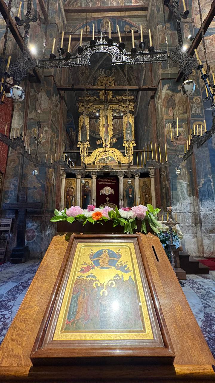 Ornate interior of an Orthodox church with gilded iconostasis, candles and fresh flowers on a wooden stand.
