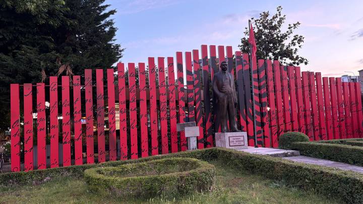 Red-and-black commemorative fence with Albanian flag motif and a statue of a national hero at dusk.