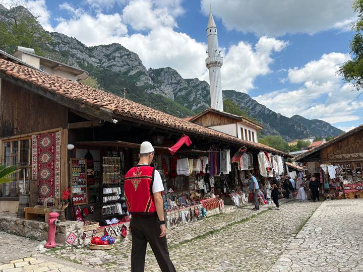 Bustling bazaar street lined with handicraft stalls beneath a white minaret and rugged mountains.