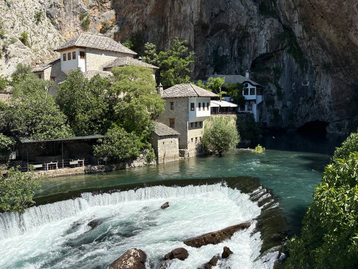 Historic Dervish monastery and stone houses beside turquoise river emerging from a cliff spring.
