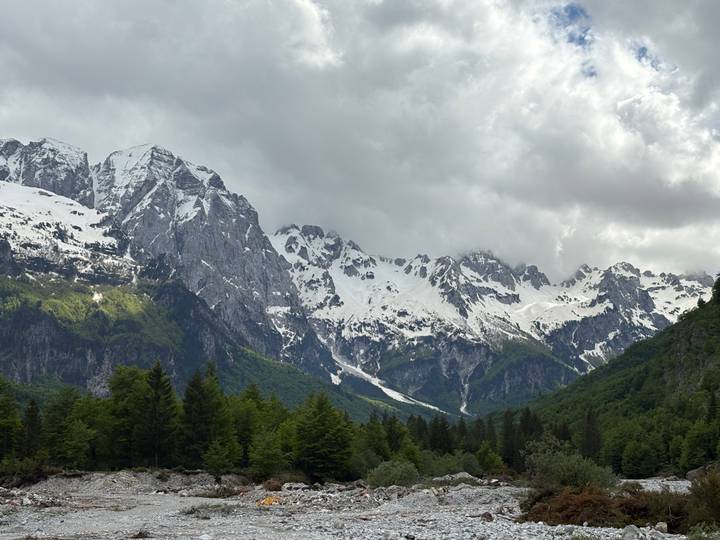 Snow-capped peaks of the Albanian Alps towering over dense green forest under dramatic clouds.