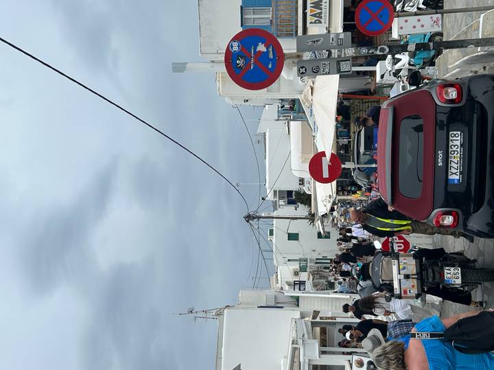 Busy street in Mykonos town filled with scooters, cars, pedestrians and white Cycladic buildings under an overcast sky.