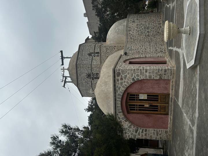 Small stone church with domes and timber doors beneath cloudy skies in Santorini.