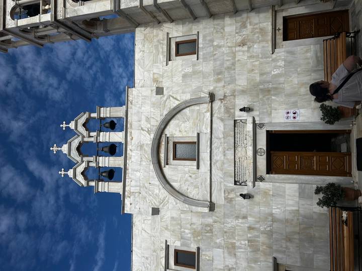 White marble church façade with triple bell tower against a vivid blue sky, visitors at entrance.