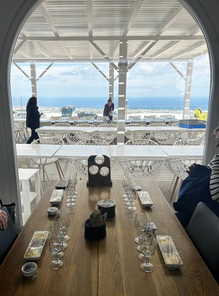 Indoor wine-tasting venue with rows of white chairs and panoramic sea view through large windows.