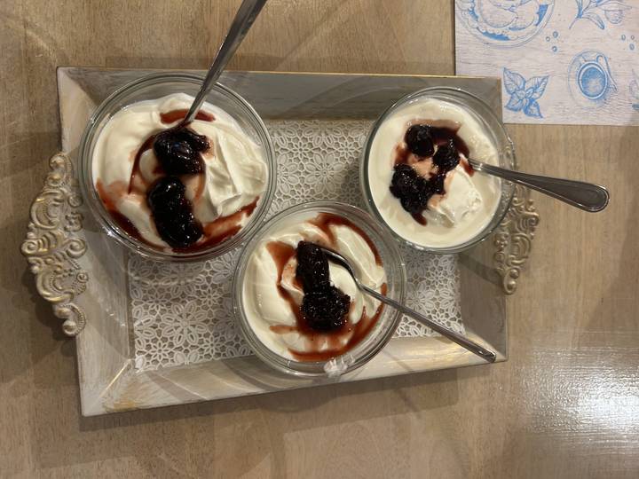 Three glass bowls of thick Greek yogurt topped with dark cherry sauce and spoons, served on a decorative wooden tray on a table.