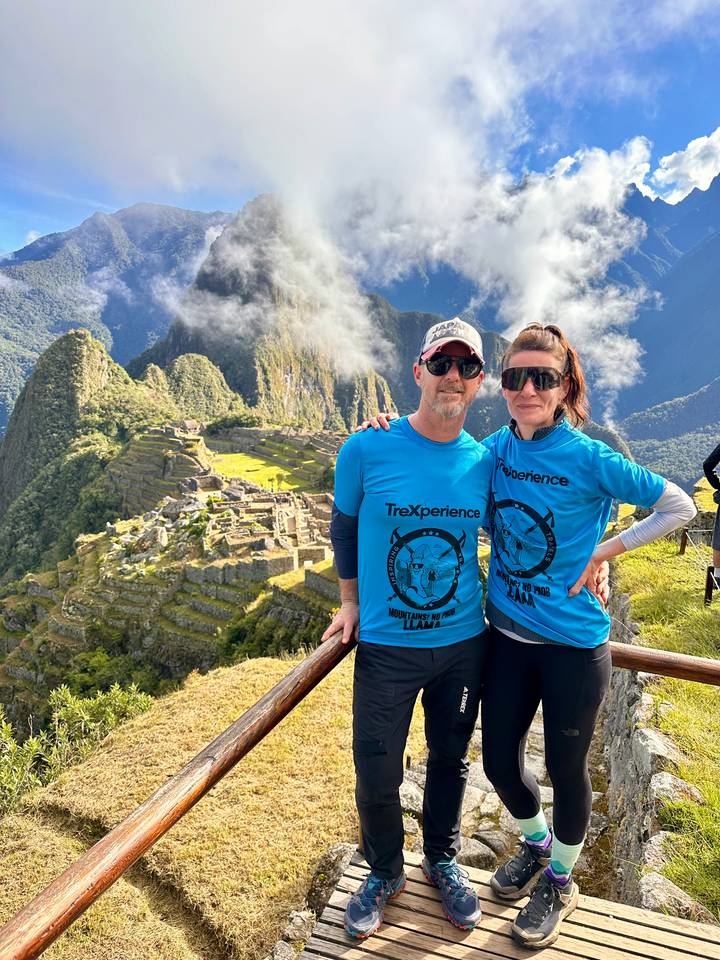 A couple in matching blue trekking shirts stand at the classic lookout over the ancient terraces of Machu Picchu with cloud-capped peaks behind.