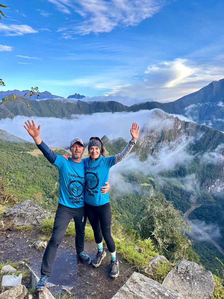 Smiling couple raise their arms triumphantly on a misty mountain overlook above lush green valleys on the Inca Trail.