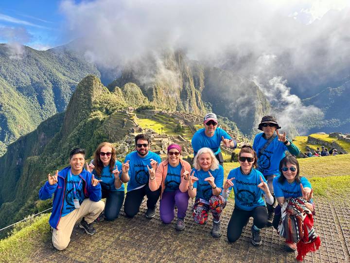Eight hikers in blue shirts flash hand signs while posing at the famous Machu Picchu lookout amid swirling clouds.