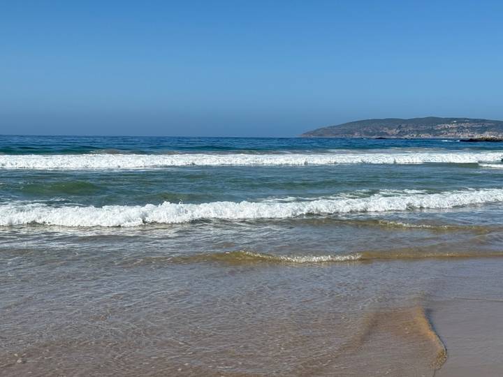 Clear turquoise waves roll onto a sandy beach with a distant green headland under a bright blue sky.