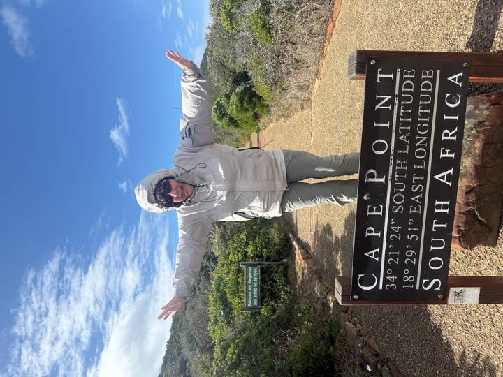 Woman in windbreaker spreads her arms behind the Cape Point latitude and longitude sign under a bright blue sky.