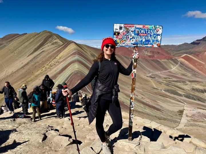 Hiker posing with a colourful sign at the summit of Rainbow Mountain with multicoloured ridges behind.