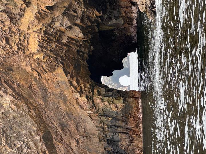 View through a rugged sea cave and rock arch toward open water, taken from a boat at water level.