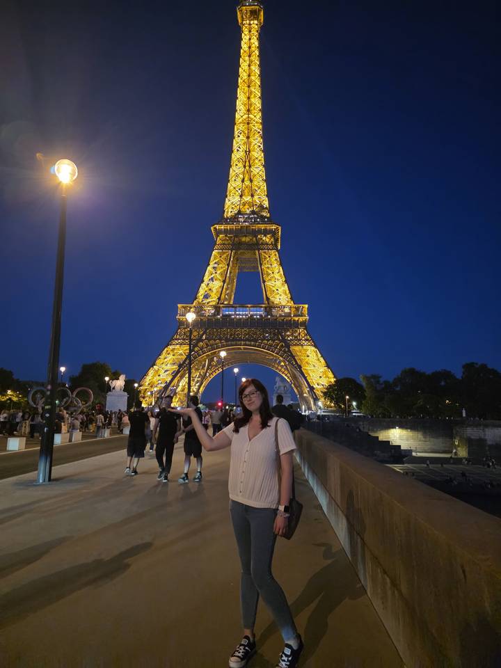 Nighttime scene of a woman posing with the illuminated Eiffel Tower and crowds in the background.