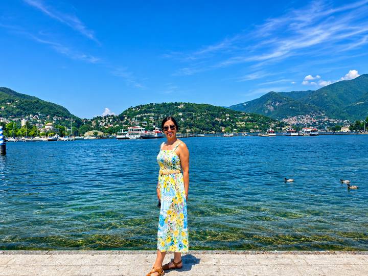 Woman in floral dress standing in clear blue lake with green hills and boats behind her.