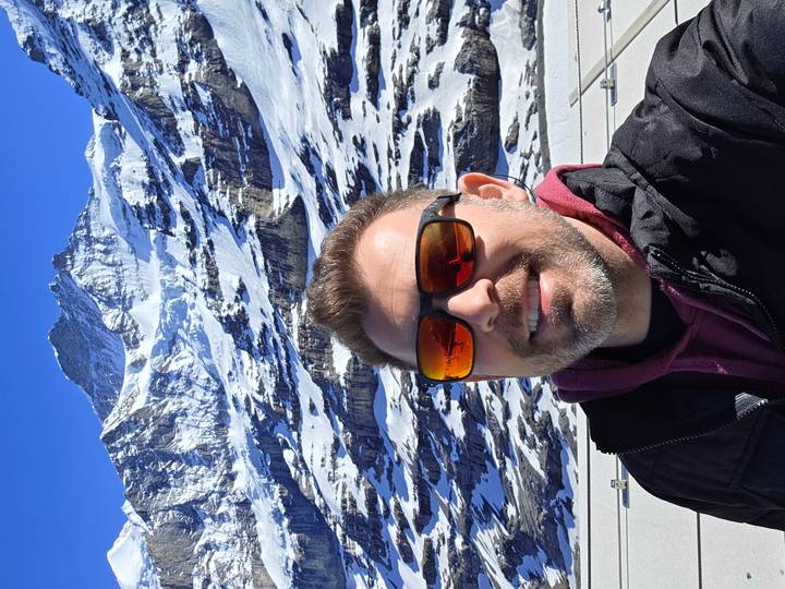 Smiling man taking a selfie with snowy alpine peaks behind him on a clear day.