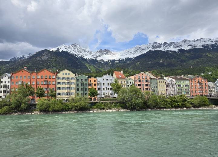 Row of colourful riverside houses with snowy Alpine mountains towering behind, reflected in turquoise water.