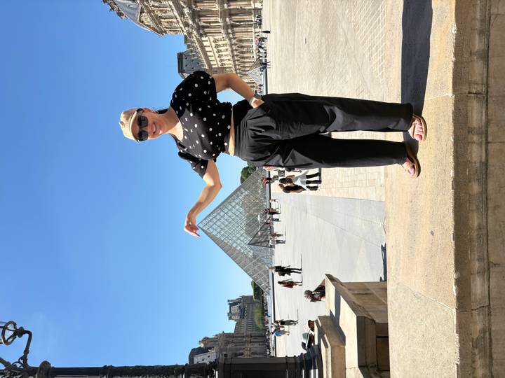 Woman mimicking touching the Louvre Pyramid while standing on a pedestal under clear skies.