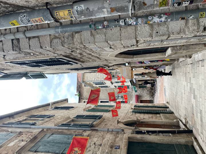 Narrow stone lane in Kotor lined with hanging Montenegrin flags and small shops.