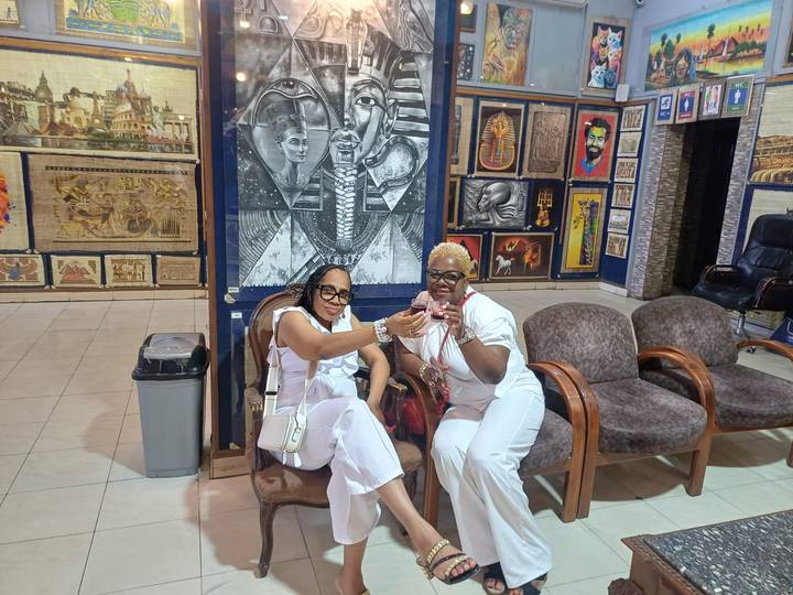 Two friends toast with drinks inside a colourful Egyptian papyrus gallery.