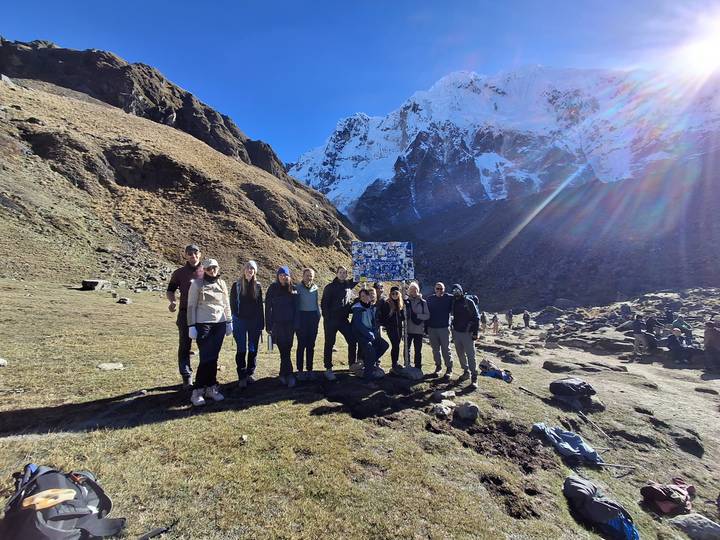 Hiking group poses triumphantly on a grassy mountain saddle beneath snow-covered Andean peaks and blue sky.