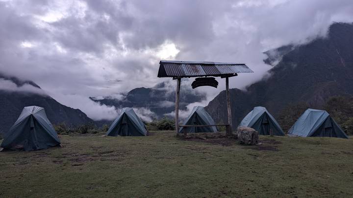 Row of green trekking tents pitched on a grassy ridge with misty cloud-filled valleys and dark mountains behind.