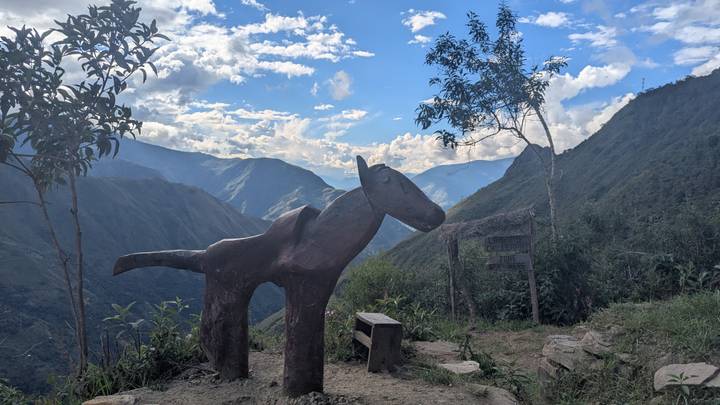 Carved wooden horse statue overlooks a dramatic Andean valley under a sky dotted with white clouds.