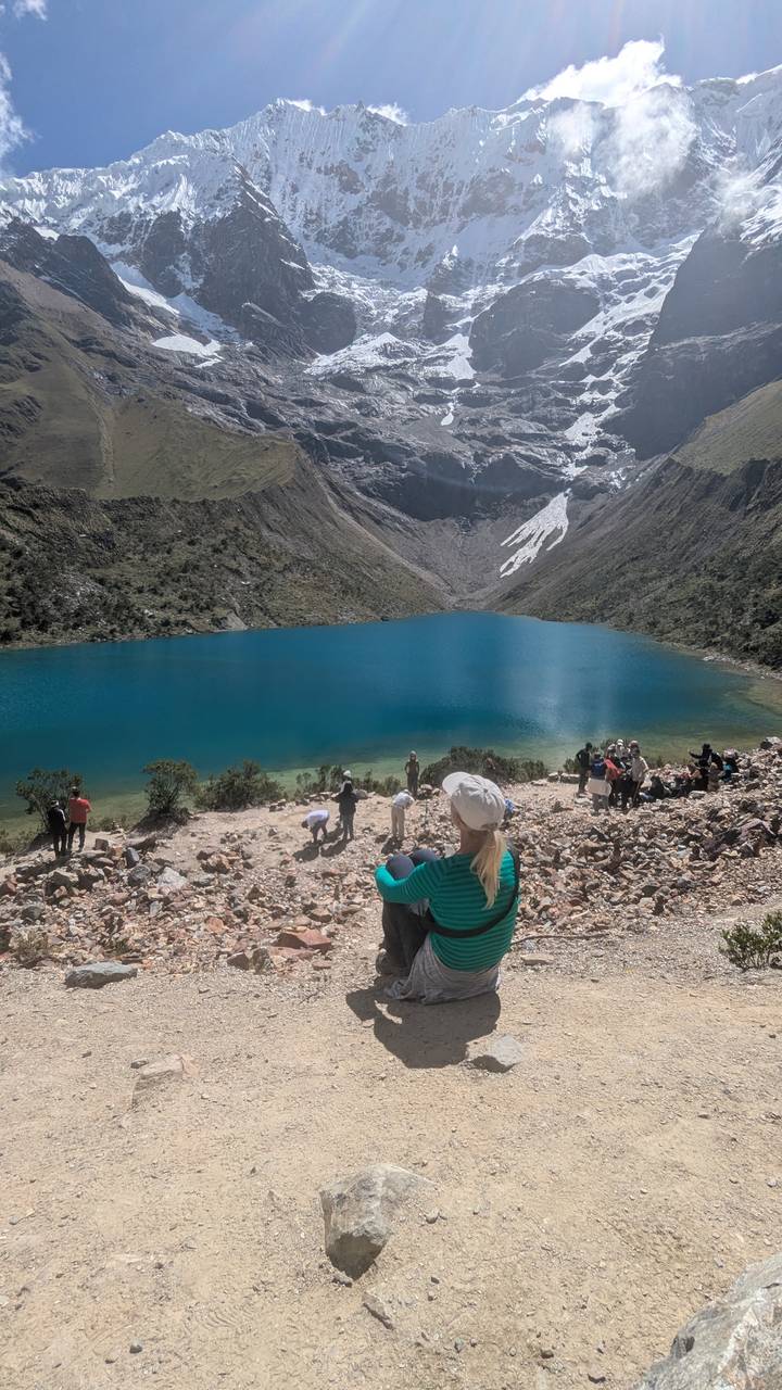Vivid turquoise glacial lake ringed by steep rocky peaks with trekkers gathered along the stony shore.