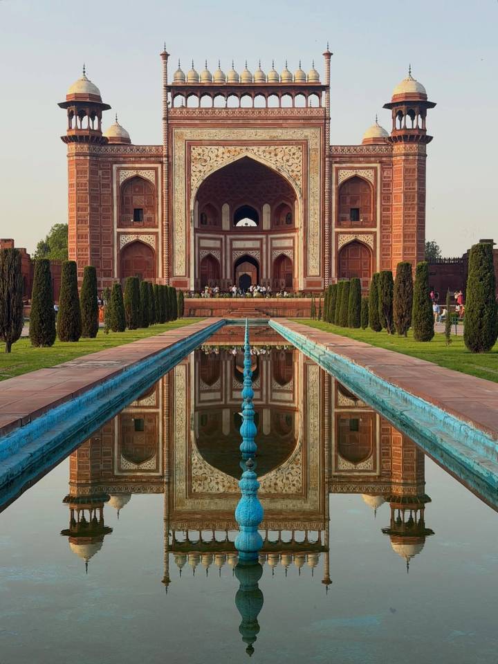 Symmetrical reflection of the grand red-sandstone gateway to the Taj Mahal complex in a long pool at sunrise.