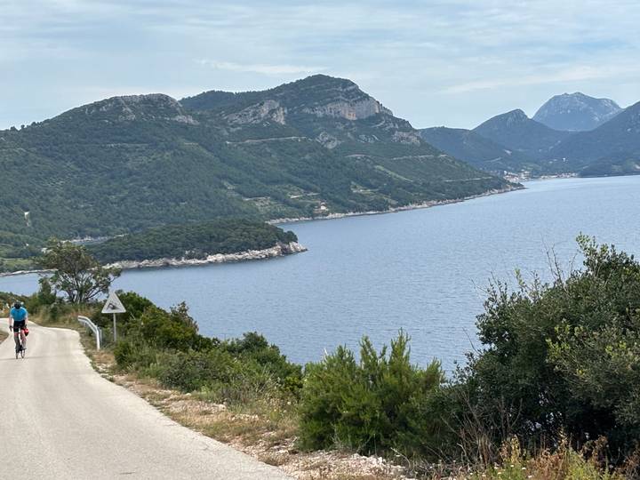 Cyclist descends quiet coastal road overlooking deep blue Adriatic coves and forested hills.